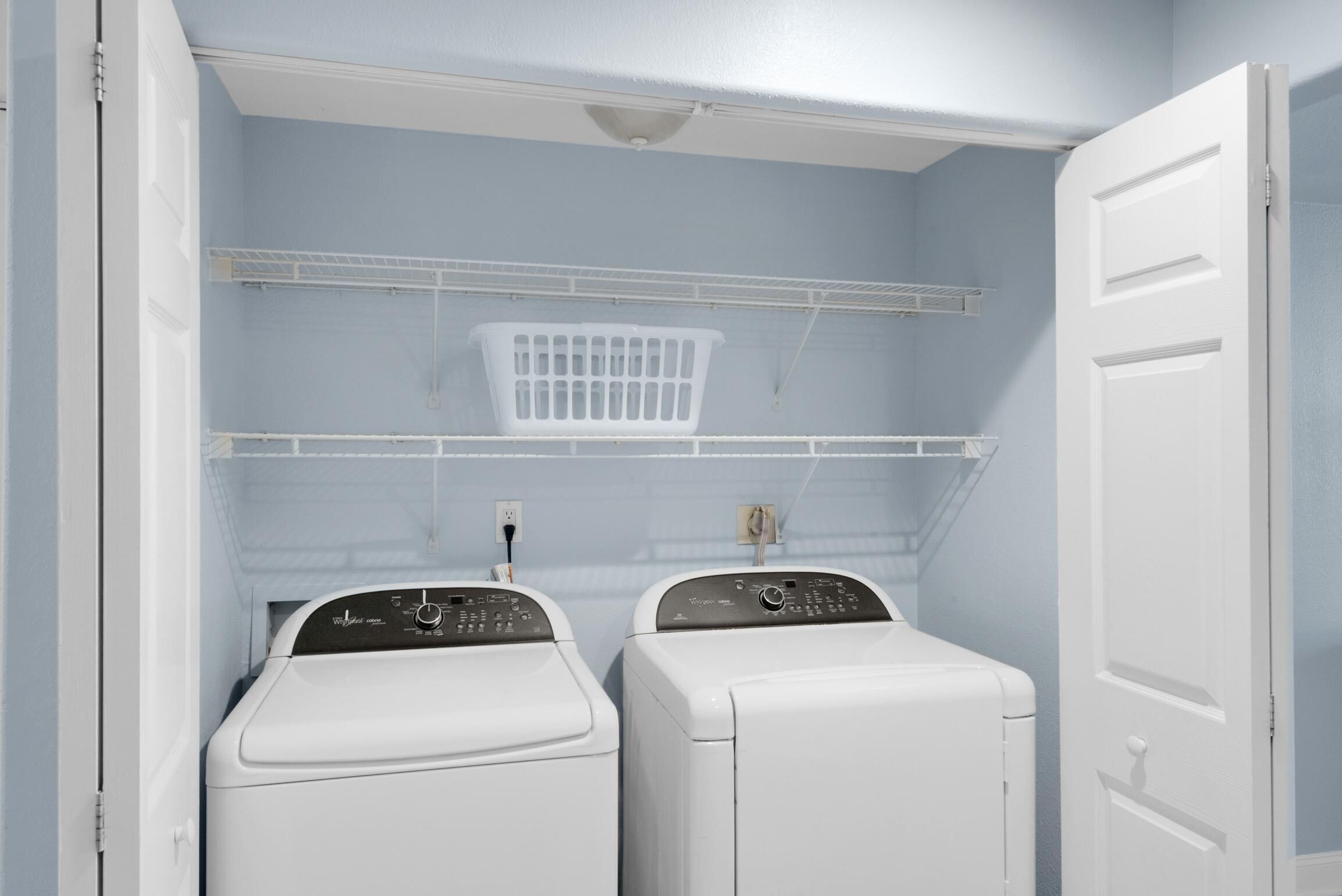 Une buanderie avec deux machines à laver et deux sèche-linge blancs, un panier à linge blanc posé sur une étagère et une porte blanche.