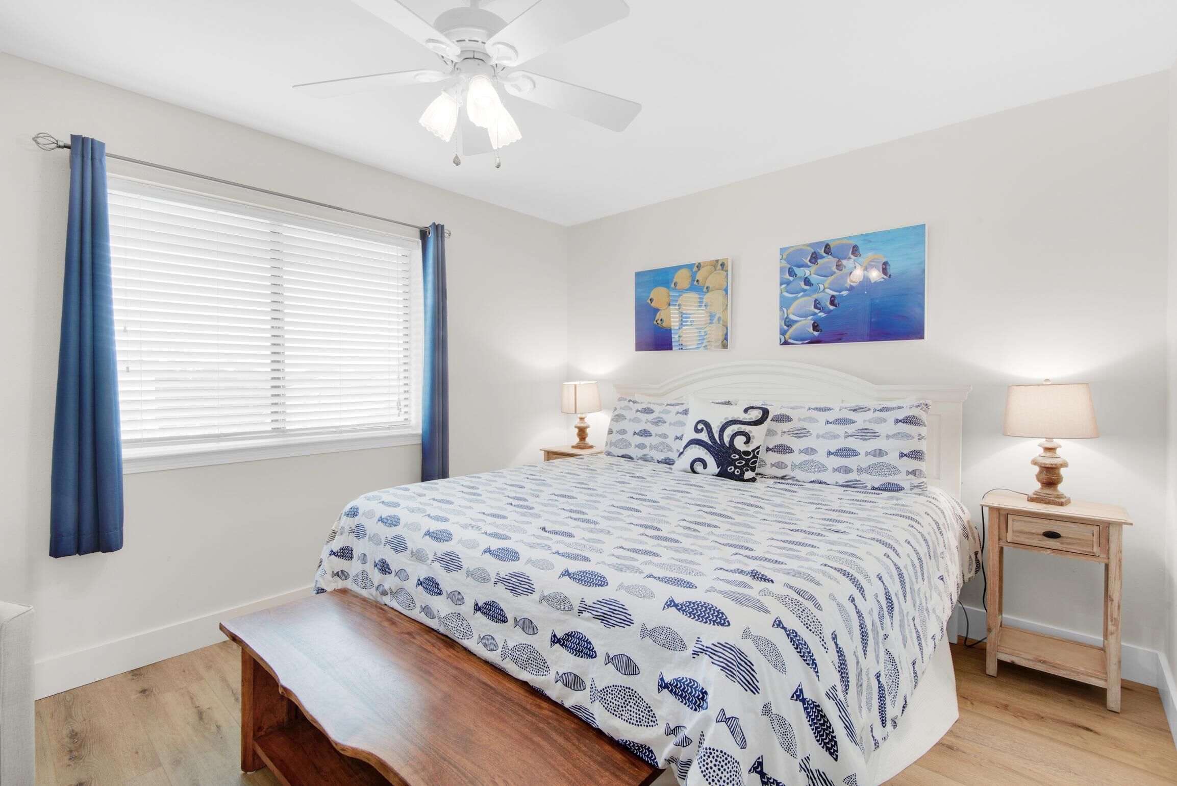 A bedroom with a bed, a ceiling fan, a window with curtains, two wall art pieces, a bedside table with a lamp, and a bench at the foot of the bed.