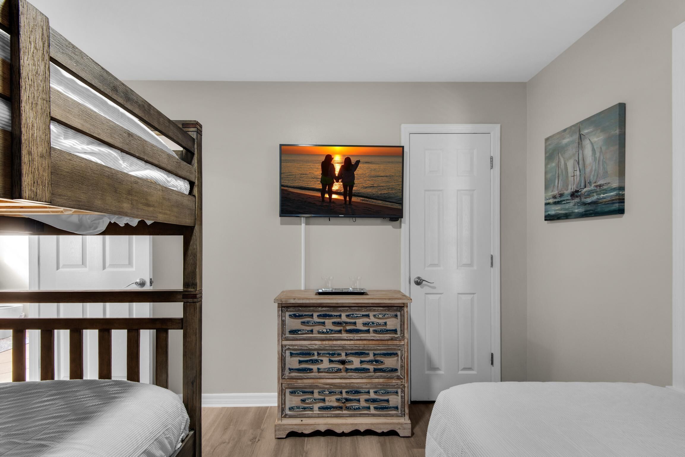 A bedroom with a bunk bed, a dresser with drawers, a television mounted on the wall, and a painting of a sailboat.