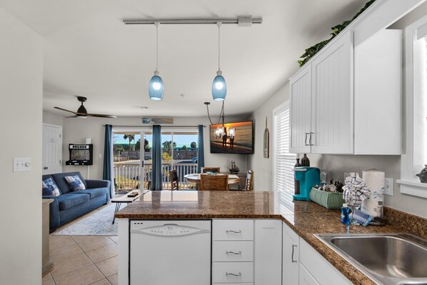 A modern kitchen with granite countertops, white cabinetry, and a stainless steel sink. There is a dining area with a table and chairs, and a balcony with a view.