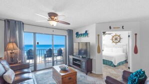 A room with a large sliding glass door to the beach, a ceiling fan, a bed with a nautical theme, and a TV.