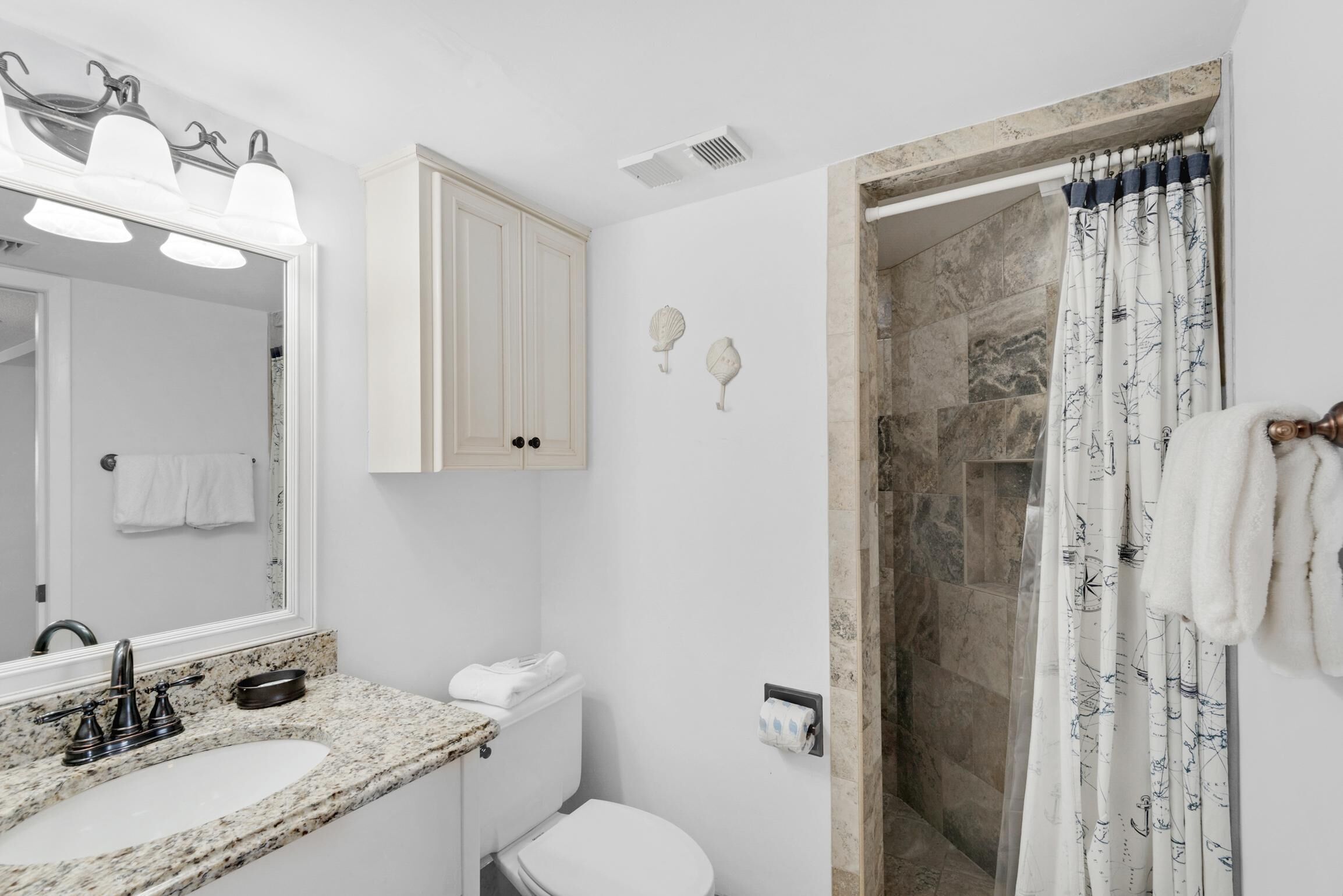 Un baño con lavamanos de encimera de granito, un inodoro blanco, un espejo con tres luces, una ducha con cortina estampada y un perchero con toallas.