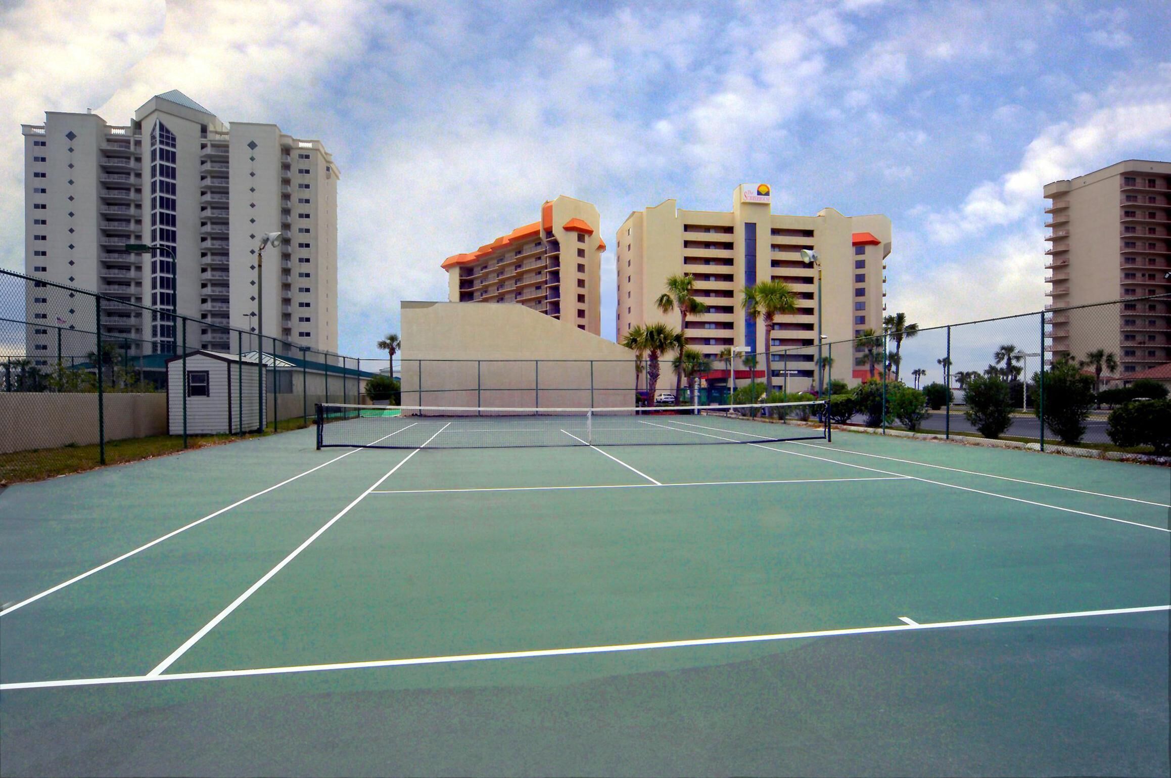 Una cancha de tenis con red, rodeada por una cerca y contigua a edificios altos.