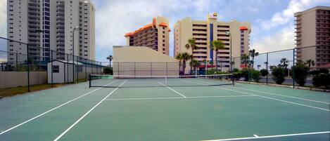 Una cancha de tenis con red, rodeada por una cerca y contigua a edificios altos.