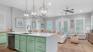 A modern kitchen with green cabinets, a white island, and pendant lights. There is a ceiling fan and a dining area with a white sofa and chairs. - Dock Holiday (Panama City Beach)