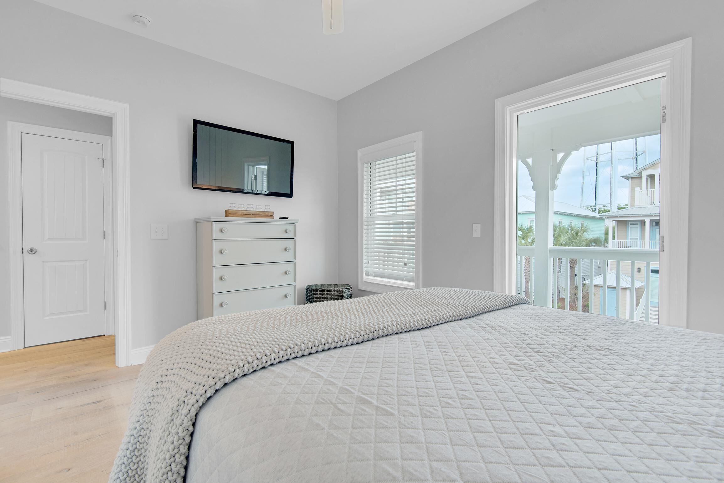 A bedroom with a large bed, a dresser, a TV mounted on the wall, and a balcony with a view of houses.