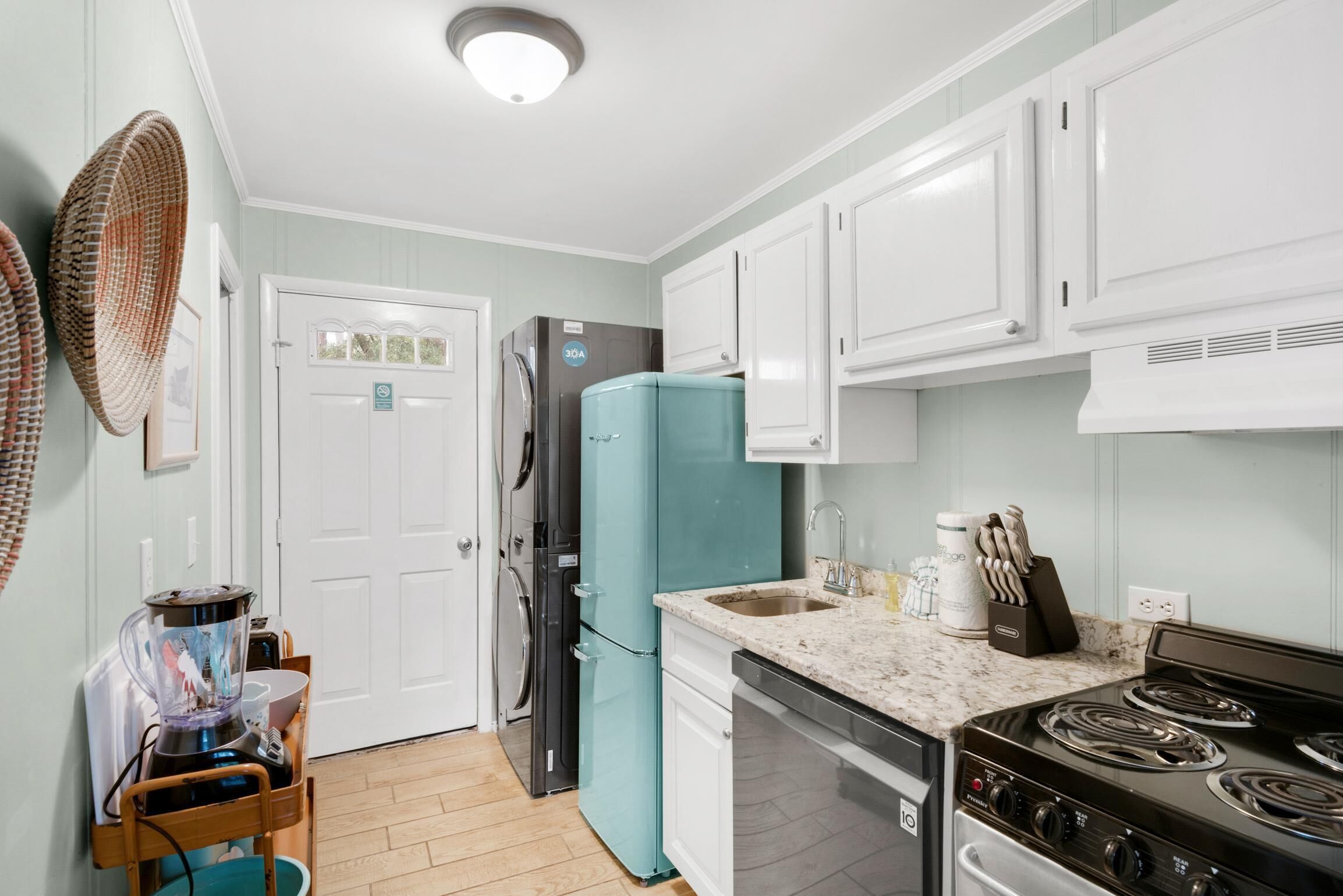 Una cocina con armarios blancos, un refrigerador verde azulado, una estufa y una licuadora sobre una silla de madera.