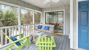Eine überdachte Veranda mit Deckenventilator, einer Holzbank mit blauen Kissen, einem grünen Adirondack-Sessel und einer Glastür, die zu einem Innenbereich führt.