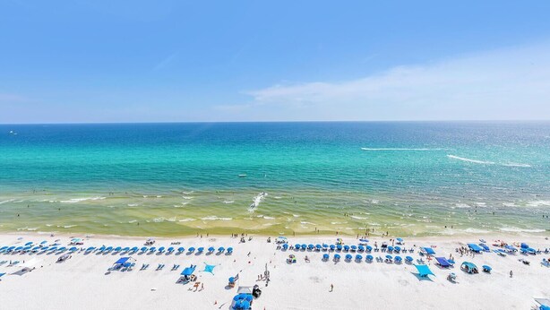 A beach with rows of blue umbrellas and sun loungers, a clear blue sea, and a sandy shore.