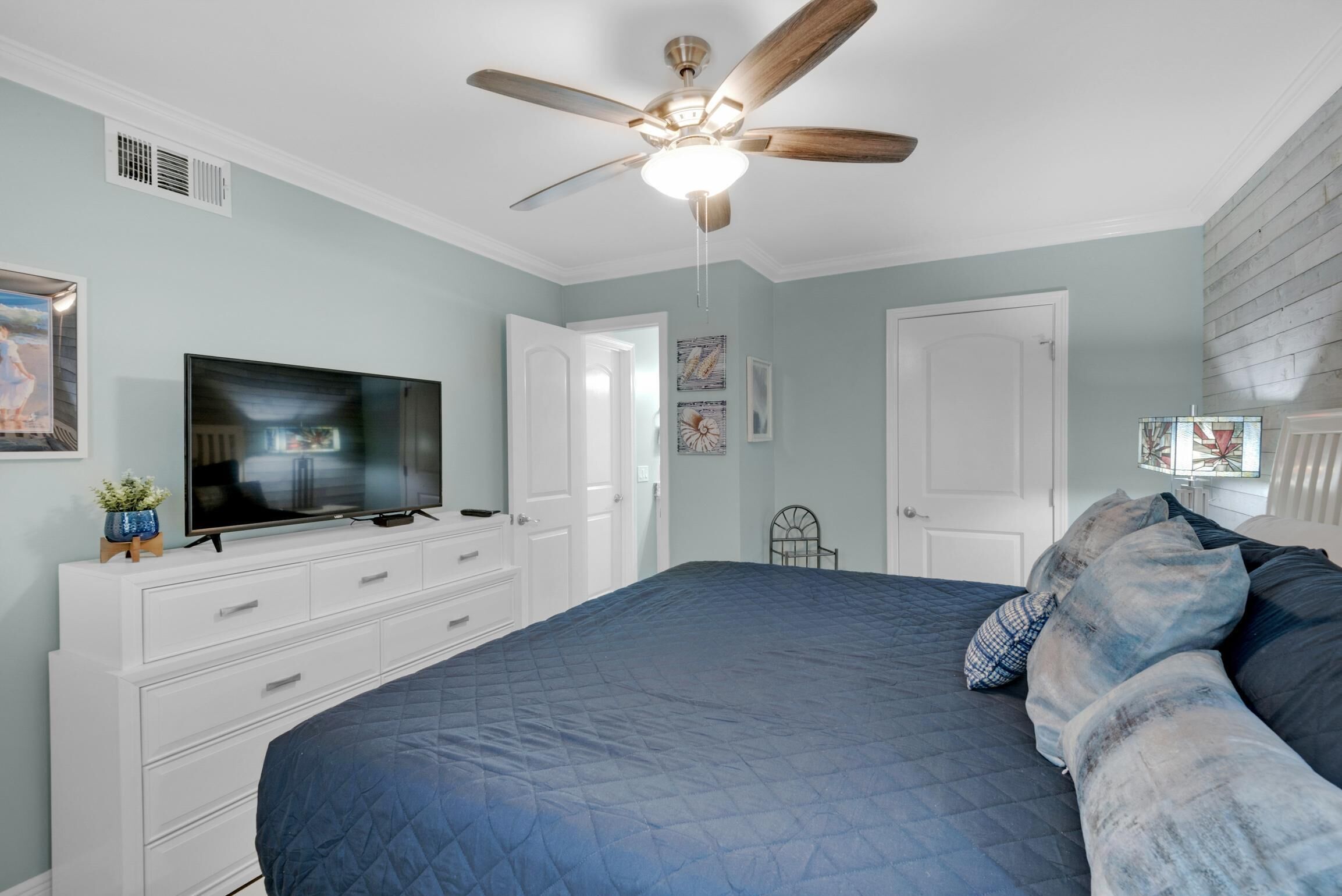 Une chambre à coucher avec un lit, un ventilateur de plafond, une télévision et des meubles blancs.