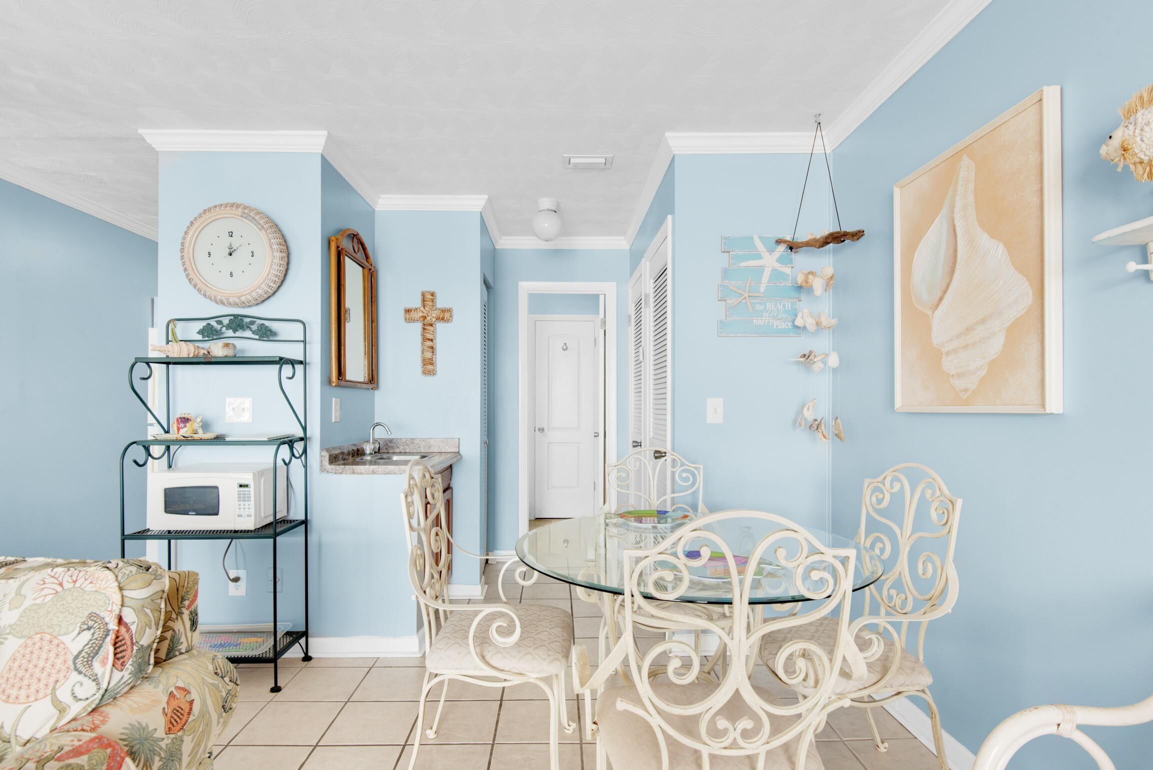 Ein Zimmer mit einem runden Glastisch und weißen Metallstühlen, einer weißen Mikrowelle auf einem Metallregal, einer Uhr an der Wand und einer Tür, die in einen anderen Raum führt.
