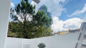 Un balcón con una silla blanca, una mesa pequeña con una planta y vistas a árboles y un edificio.