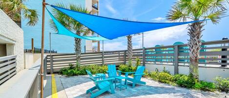 An outdoor patio with blue chairs and a table, a blue shade sail, and a white building in the background.