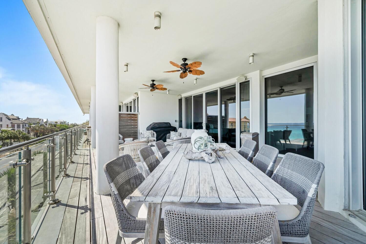 A covered outdoor dining area with a long wooden table, wicker chairs, ceiling fans, and a view of the ocean.