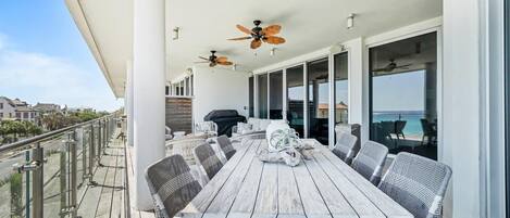 A covered outdoor dining area with a long wooden table, wicker chairs, ceiling fans, and a view of the ocean.