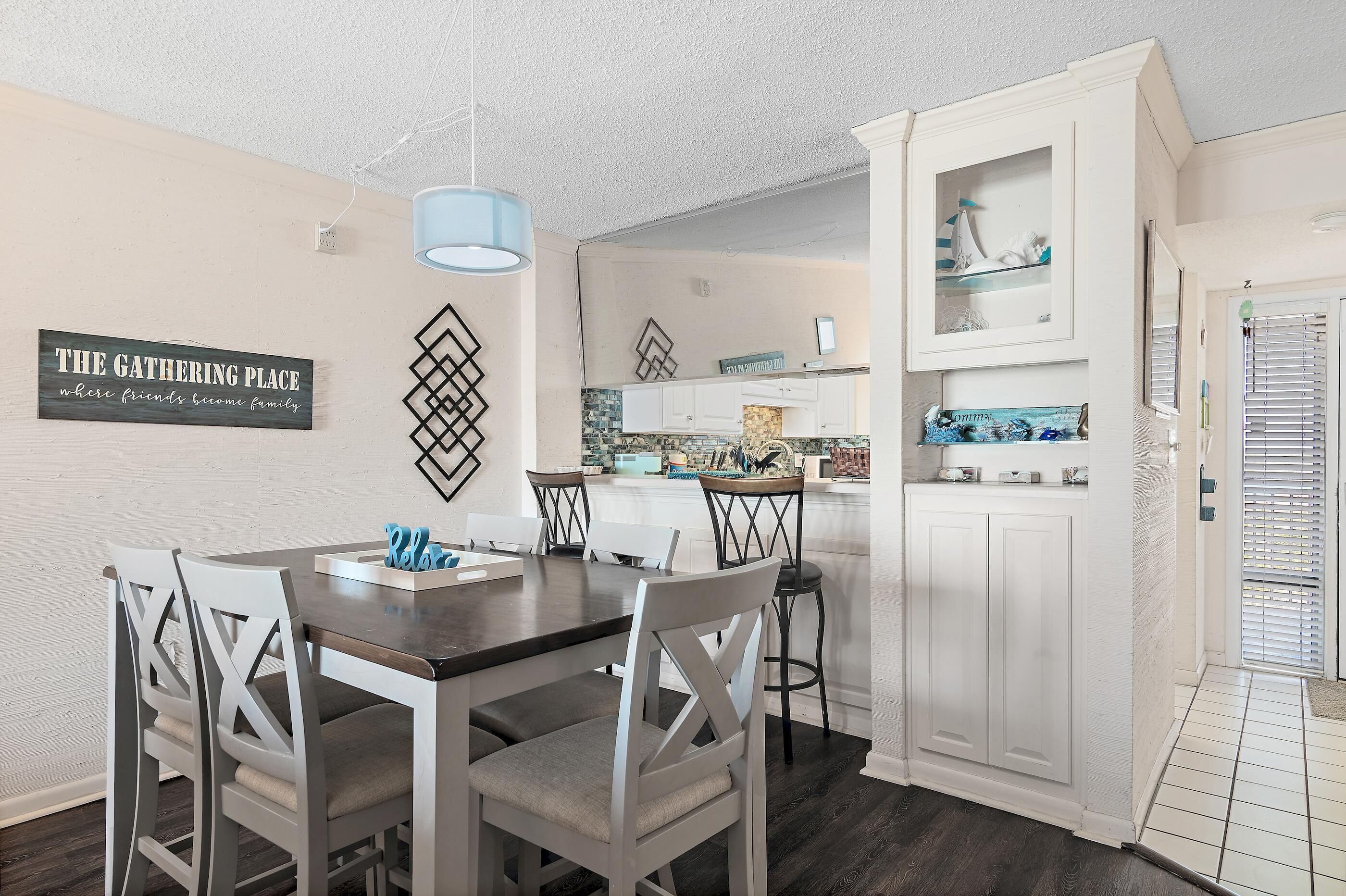A dining area with a dark wooden table and white chairs, a blue pendant light, and a sign on the wall reading "THE GATHERING PLACE where friends become family."