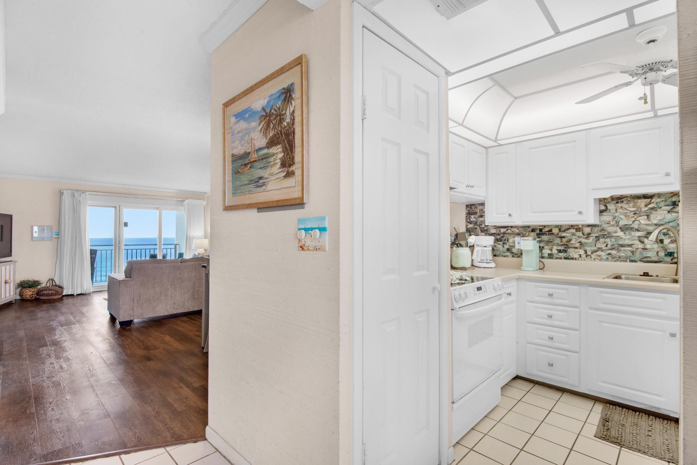 A spacious living area with a large window, a sofa, and a kitchen with white cabinets and a tiled backsplash.