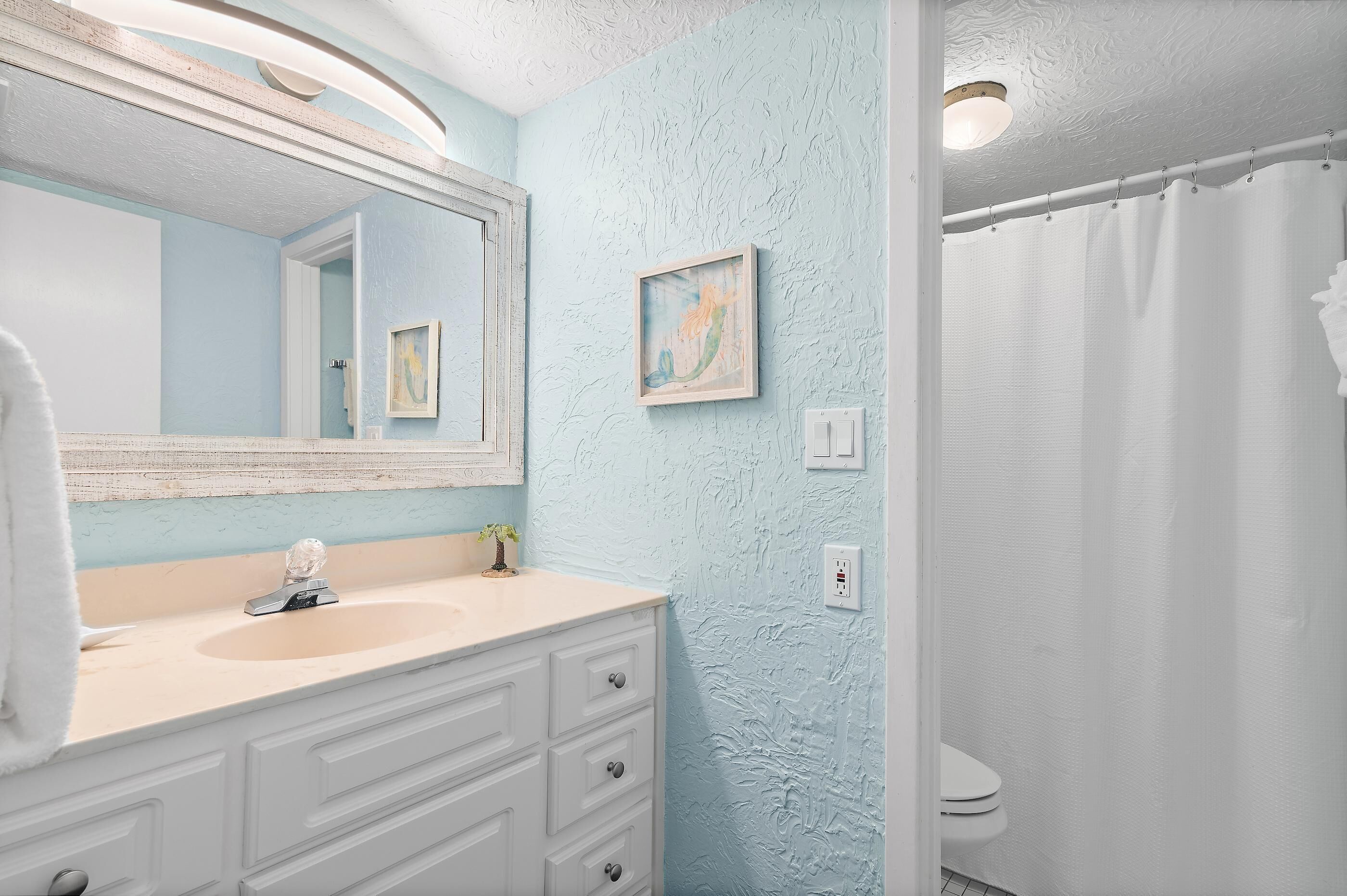 Een badkamer met een witte wastafel, een grote spiegel, een klein, ingelijst schilderij en een witte douchegordijn.