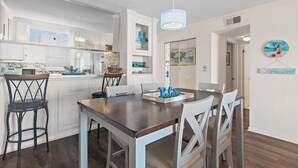A modern kitchen with white cabinetry, a dining area with a wooden table and chairs, and a pendant light. - Emerald Isle 708 (Fort Walton Beach)