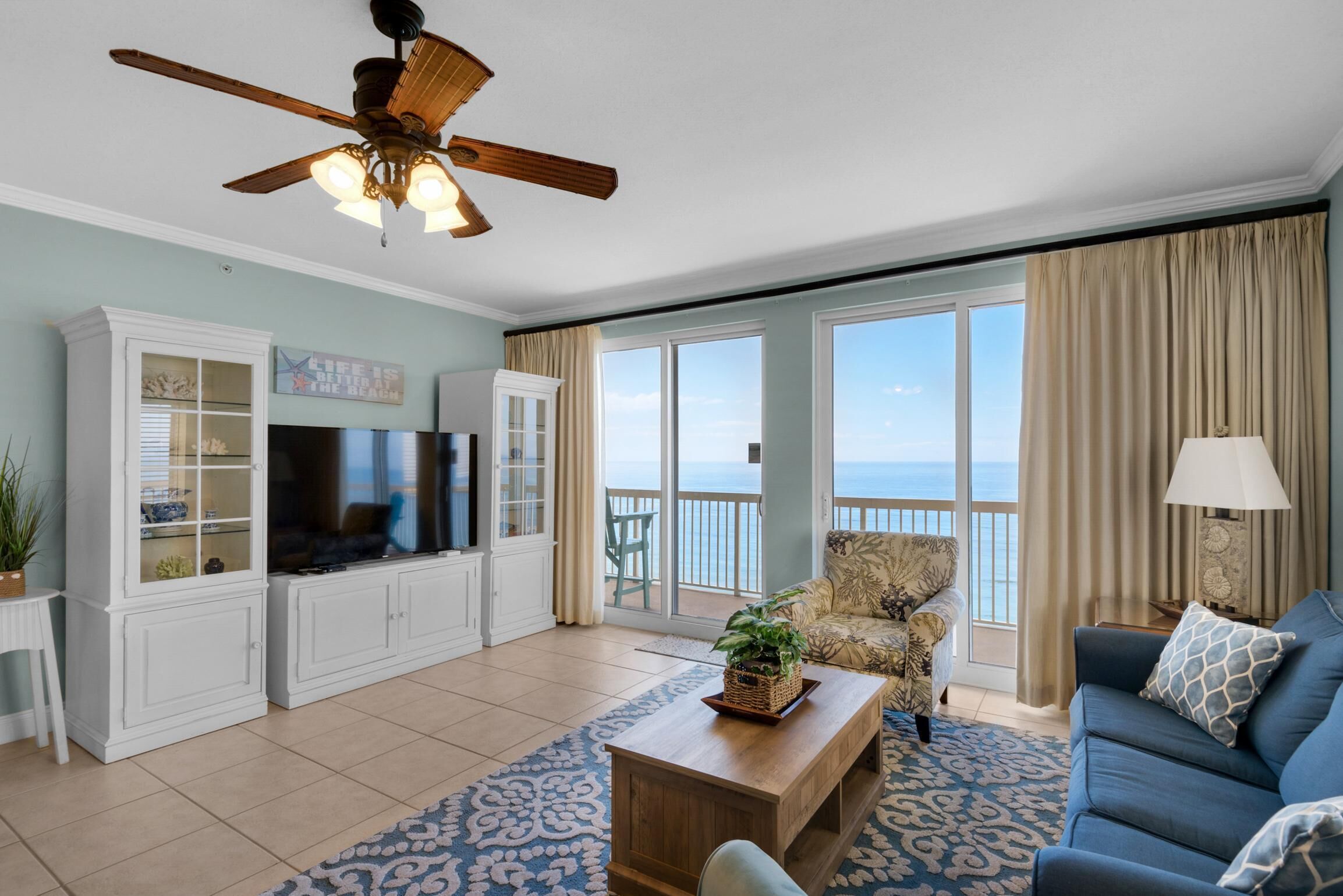 A spacious living room with a large TV, white cabinetry, a ceiling fan, a blue sofa, and a view of the ocean through large windows.