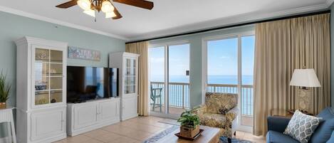 A spacious living room with a large TV, white cabinetry, a ceiling fan, a blue sofa, and a view of the ocean through large windows.