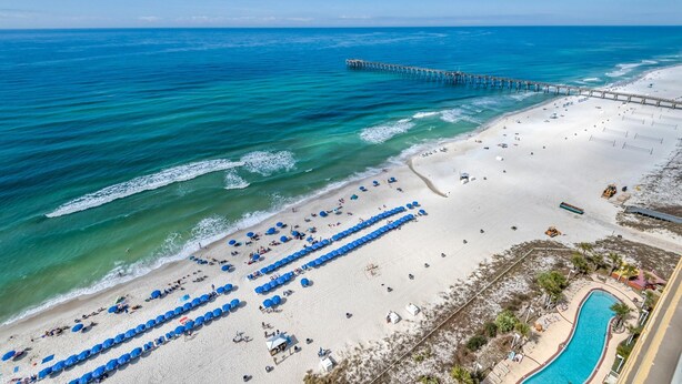 Una playa con un muelle que se adentra en el océano, una piscina y varias filas de sombrillas azules.
