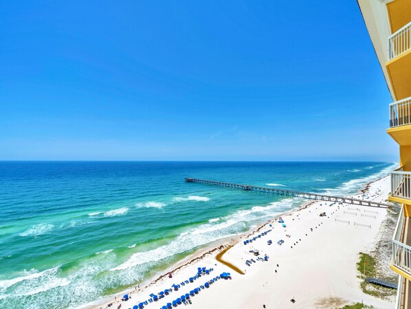 A beach with a pier extending into the ocean, a sandy beach with blue umbrellas, and a building with balconies overlooking the beach.