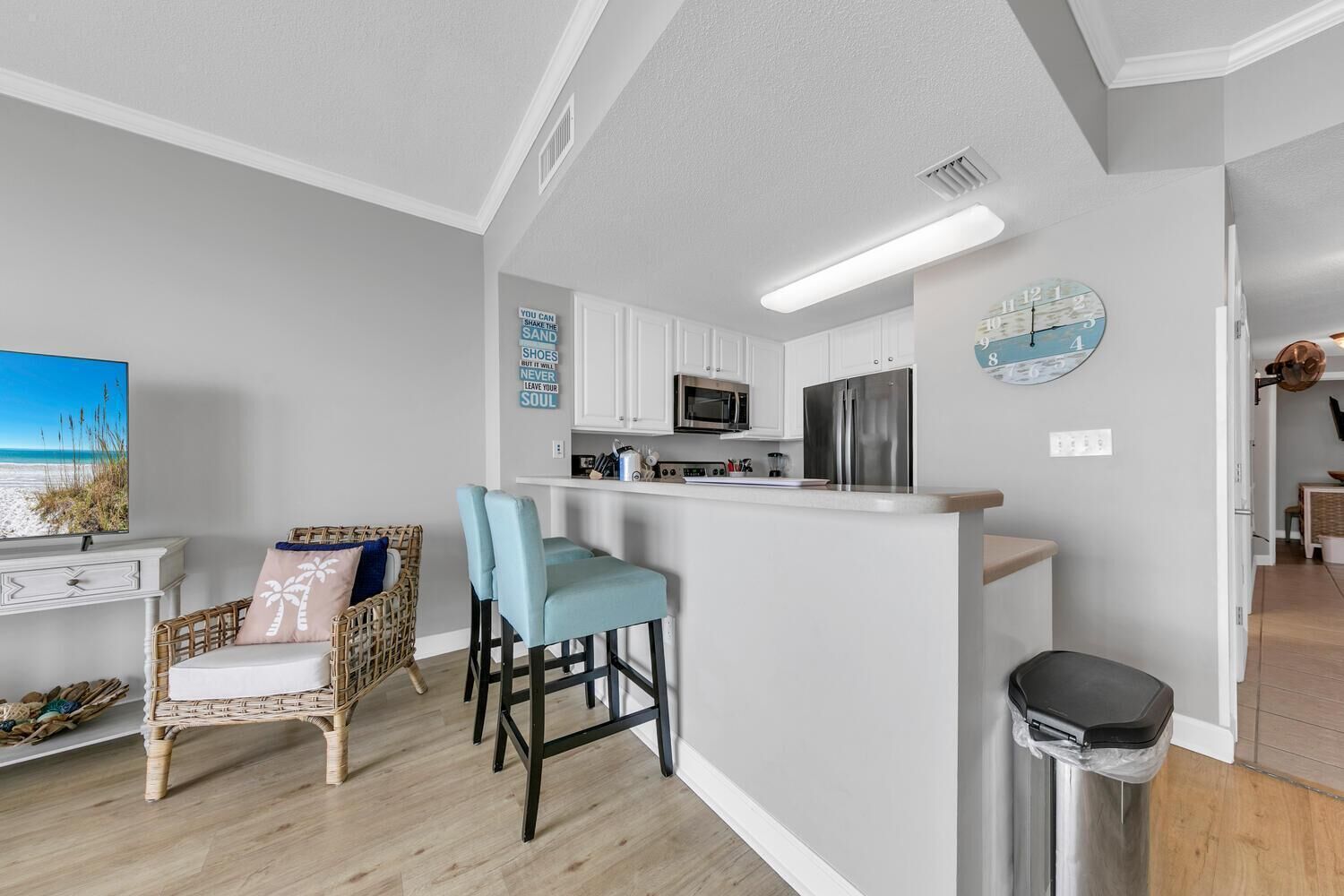 A modern kitchen with white cabinets, a stainless steel refrigerator, and a microwave. There is a bar area with two blue barstools and a wicker chair with a cushion. A trash can is visible to the right.