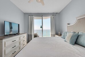 A bedroom with a large bed, a television on a dresser, and a balcony with a view of the sea.