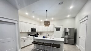 A modern kitchen with white cabinets, a stainless steel refrigerator, a central island with bar stools, and recessed ceiling lighting.