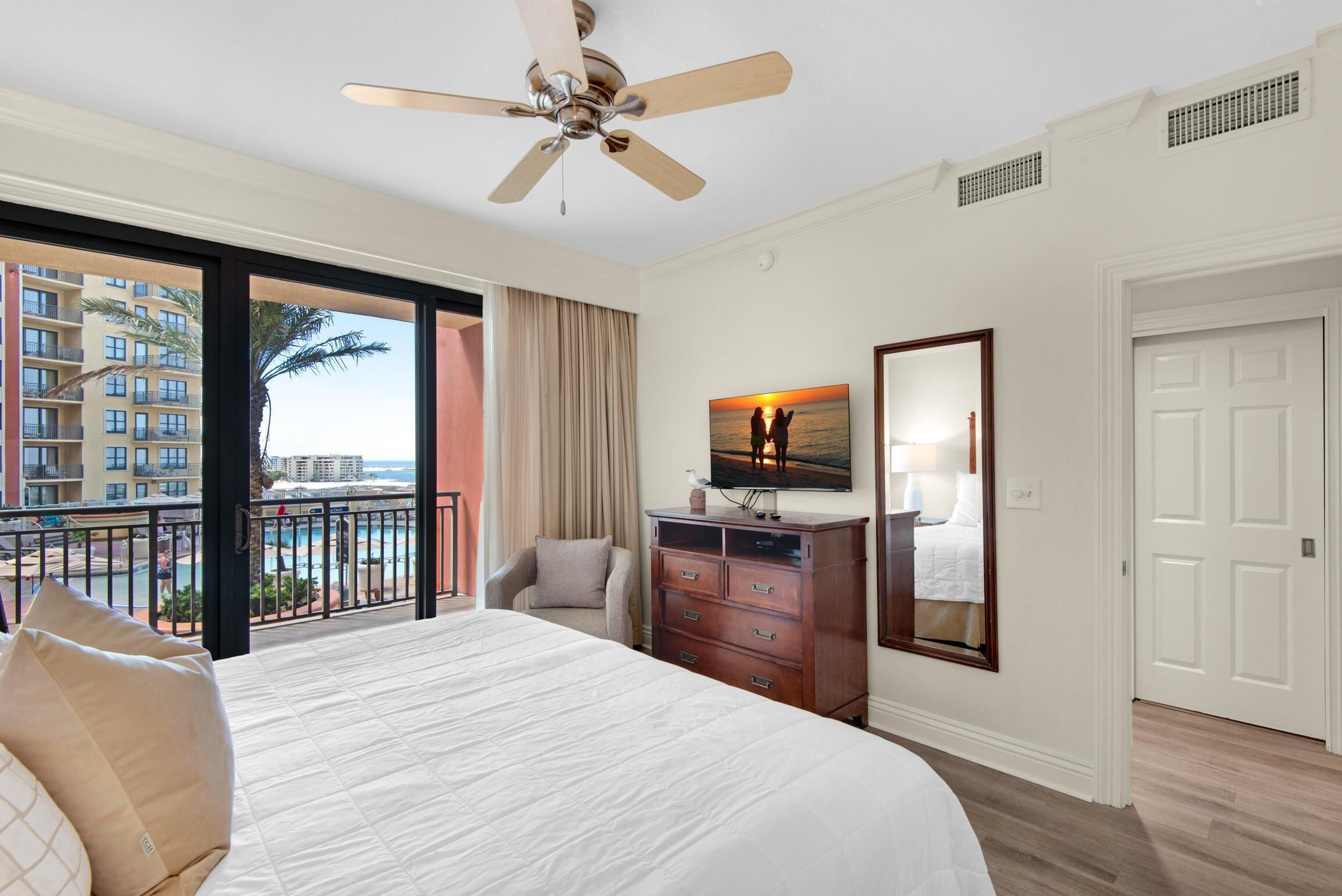 A hotel room with a large bed, a ceiling fan, a view of a building and a pool, a wooden dresser, and a door.