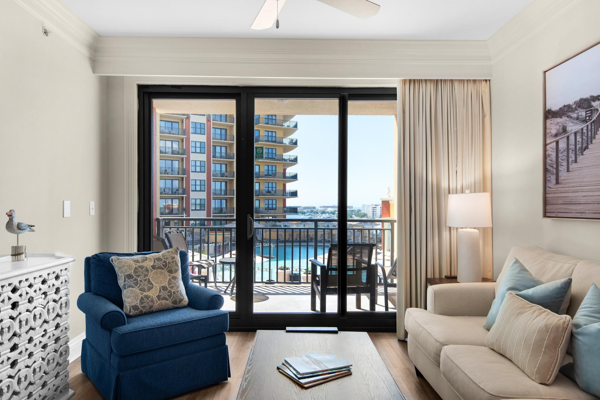 Habitación de hotel con balcón, un sillón azul, un sofá color beige, una mesa de centro de madera y vistas a un edificio alto y una piscina.
