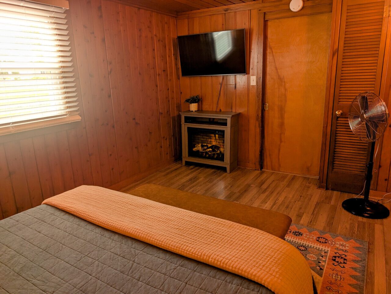 Quarto aconchegante com paredes de madeira, uma cama com colcha, uma televisão montada acima de uma lareira e um ventilador.