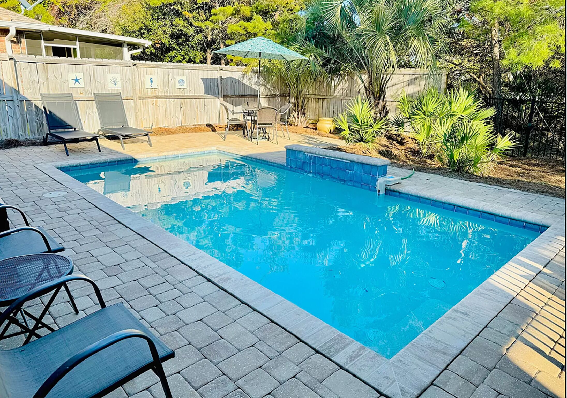 Una piscina rectangular con borde de azulejos, rodeada por un patio con sillas y una mesa.