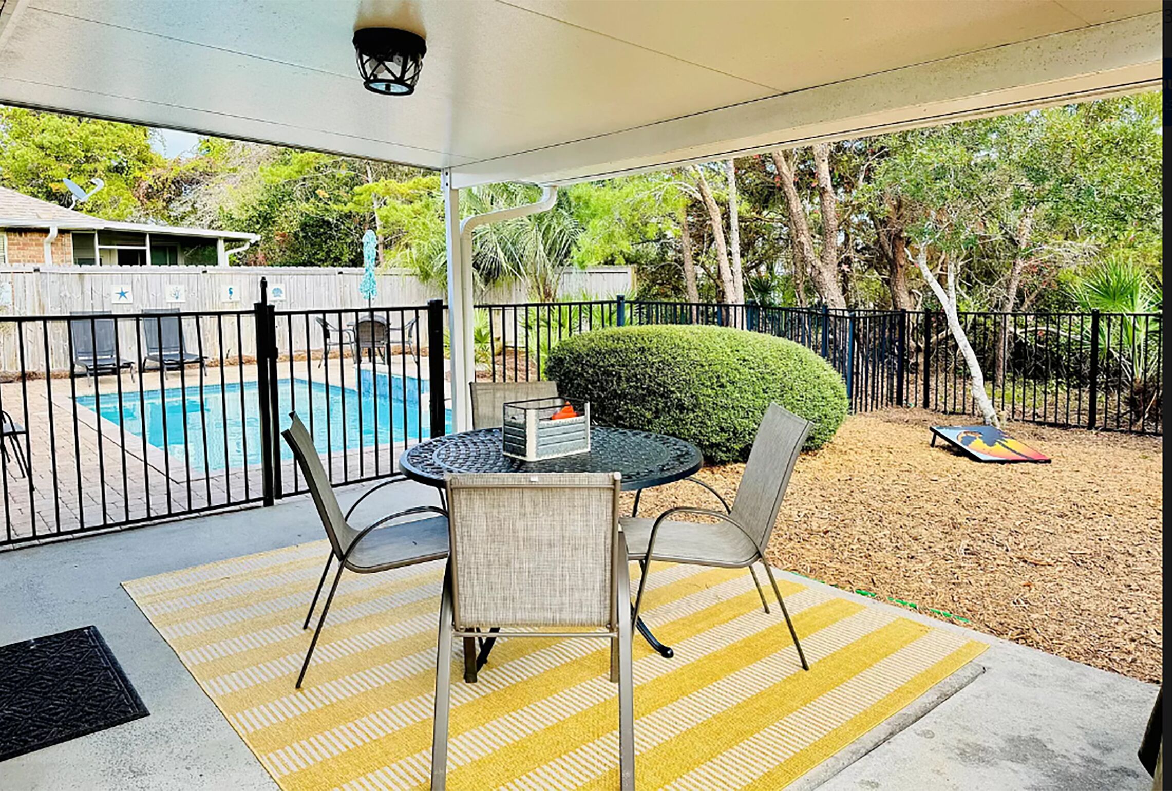 Un patio techado con una mesa y sillas, una piscina al fondo y un jardín cercado con árboles y arbustos.