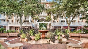 A courtyard with a central fountain, surrounded by potted plants and benches, with a building featuring balconies in the background.