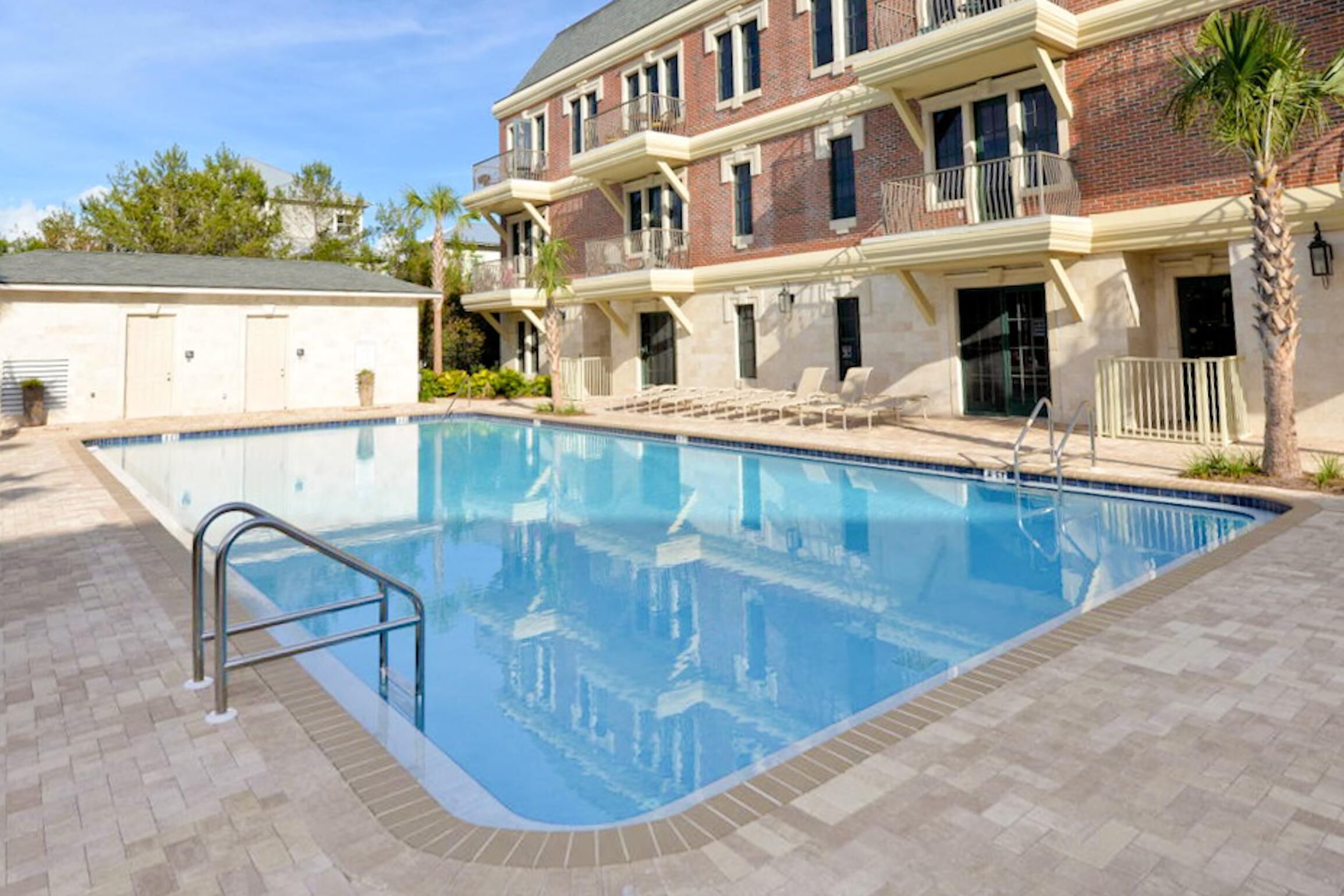 A swimming pool with a metal ladder, surrounded by a brick building with balconies and a palm tree.