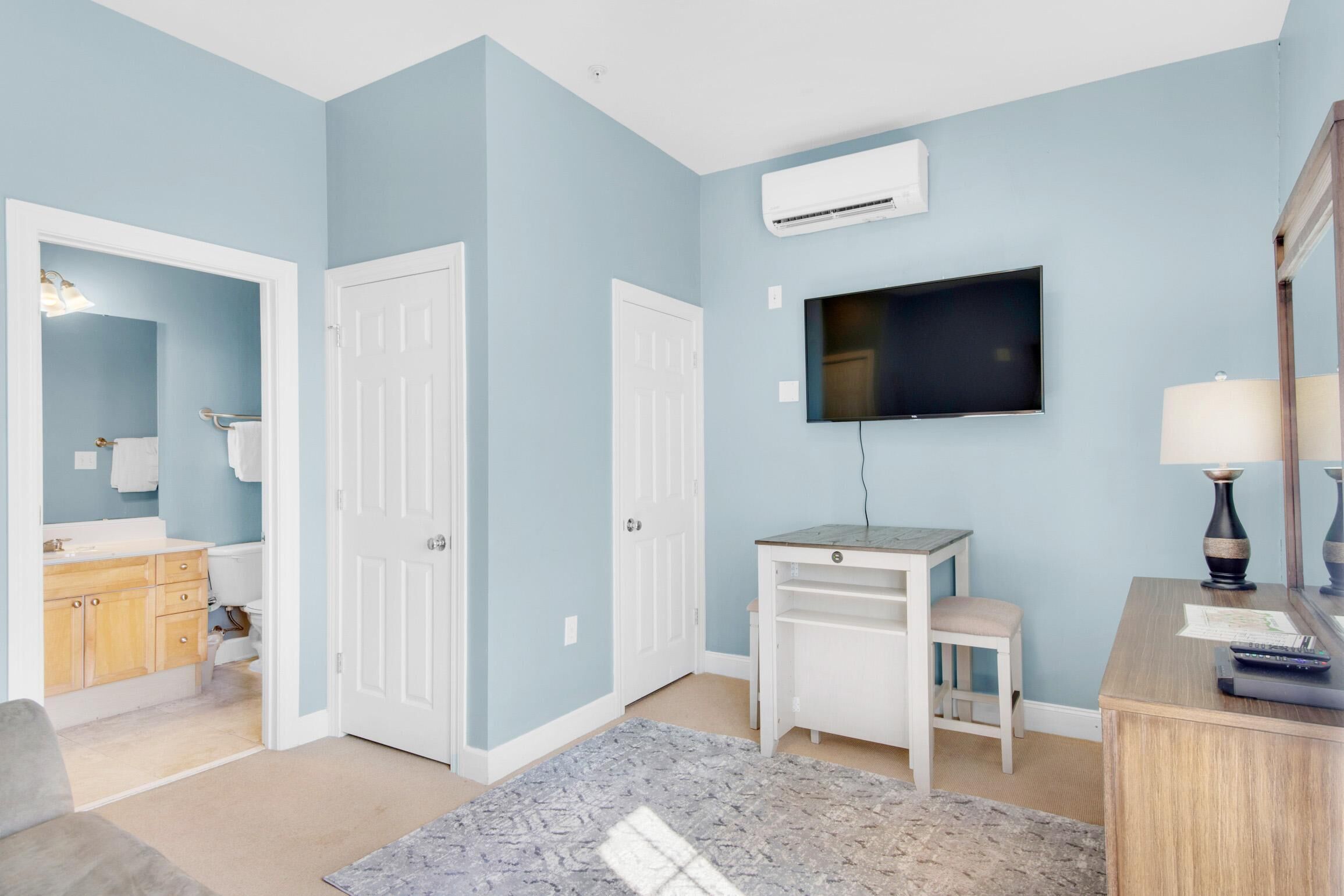 A room with light blue walls, a flat-screen TV mounted on the wall, a white desk with a lamp, and a grey rug on the floor.