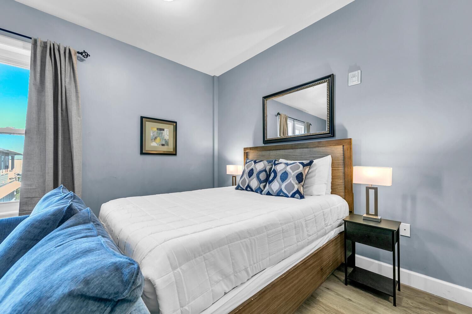 A bedroom with a bed, a nightstand, a lamp, a framed picture, and a view of the outside.