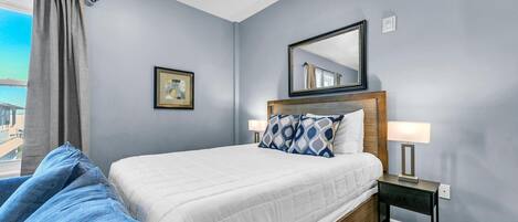 A bedroom with a bed, a nightstand, a lamp, a framed picture, and a view of the outside.