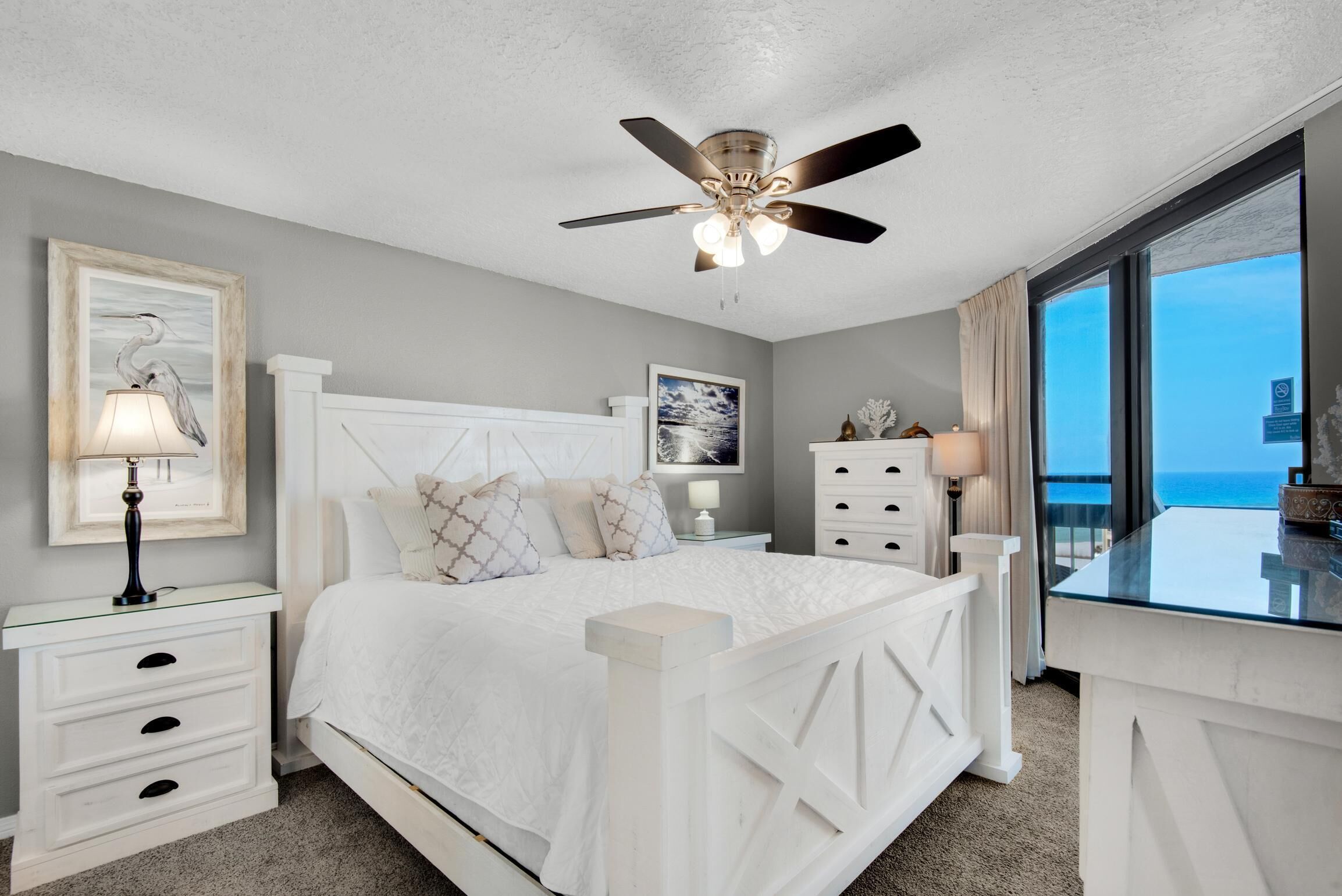 A bedroom with a bed, nightstands, a ceiling fan, a window with a view, and a framed picture on the wall.