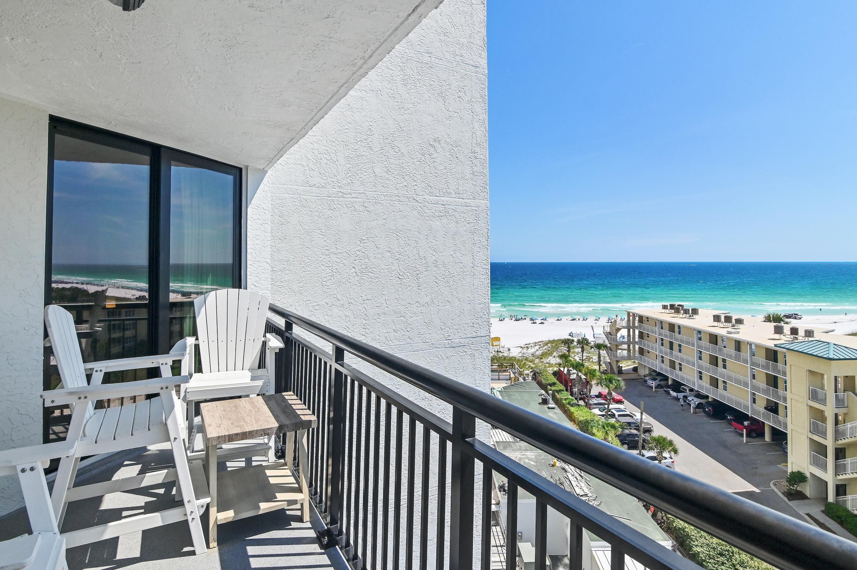 A balcony with white chairs and a table, overlooking a beach with a hotel building in the background.