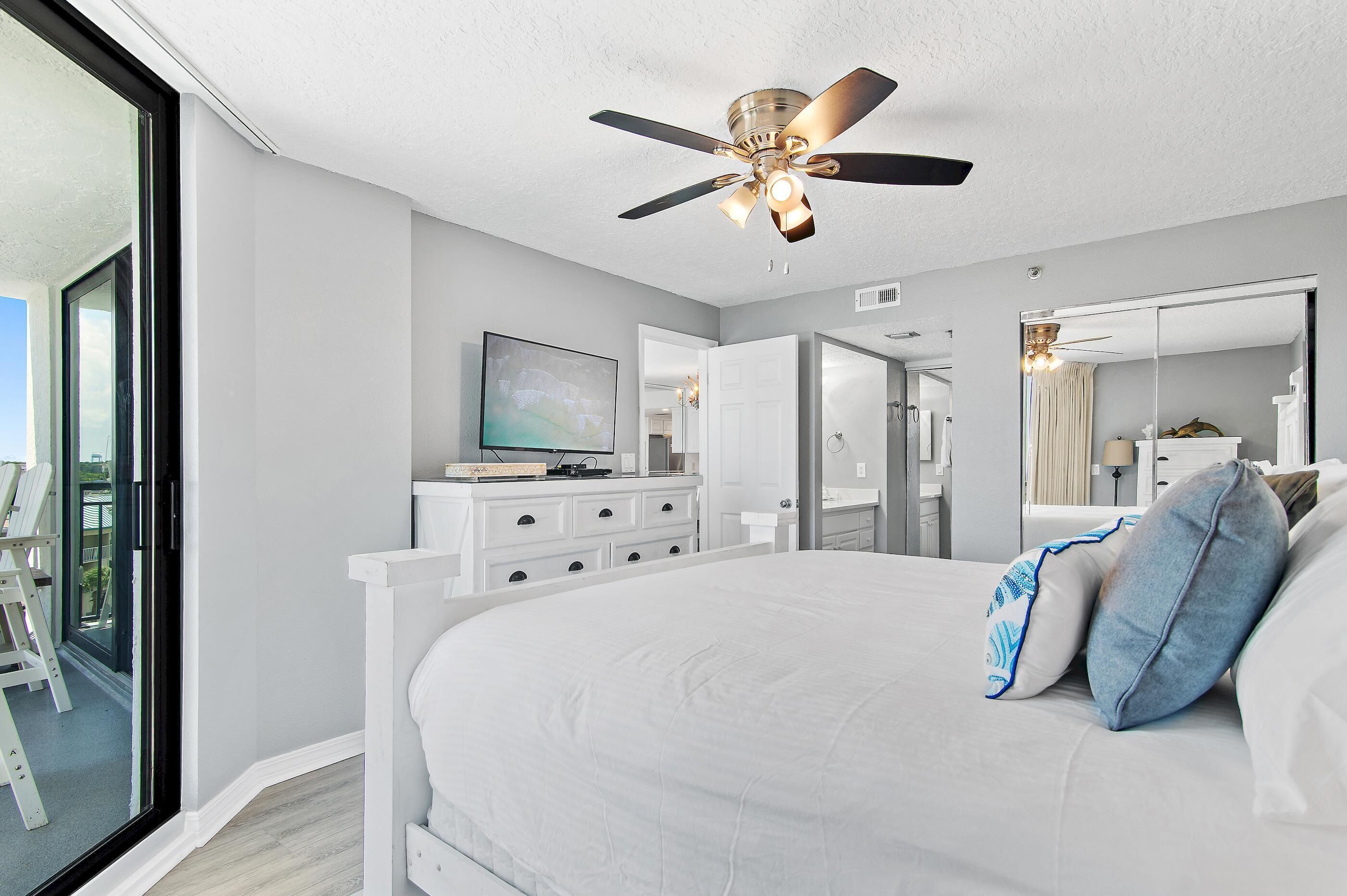 A bedroom with a large bed, a ceiling fan, a TV, and a sliding glass door to a balcony.