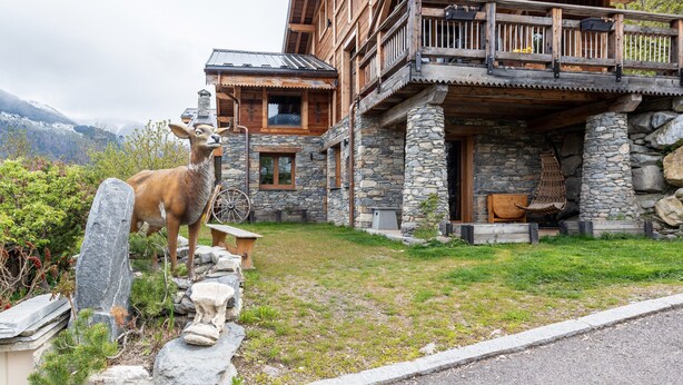 A wooden cabin with stone walls, a balcony, and a deer statue in the yard.