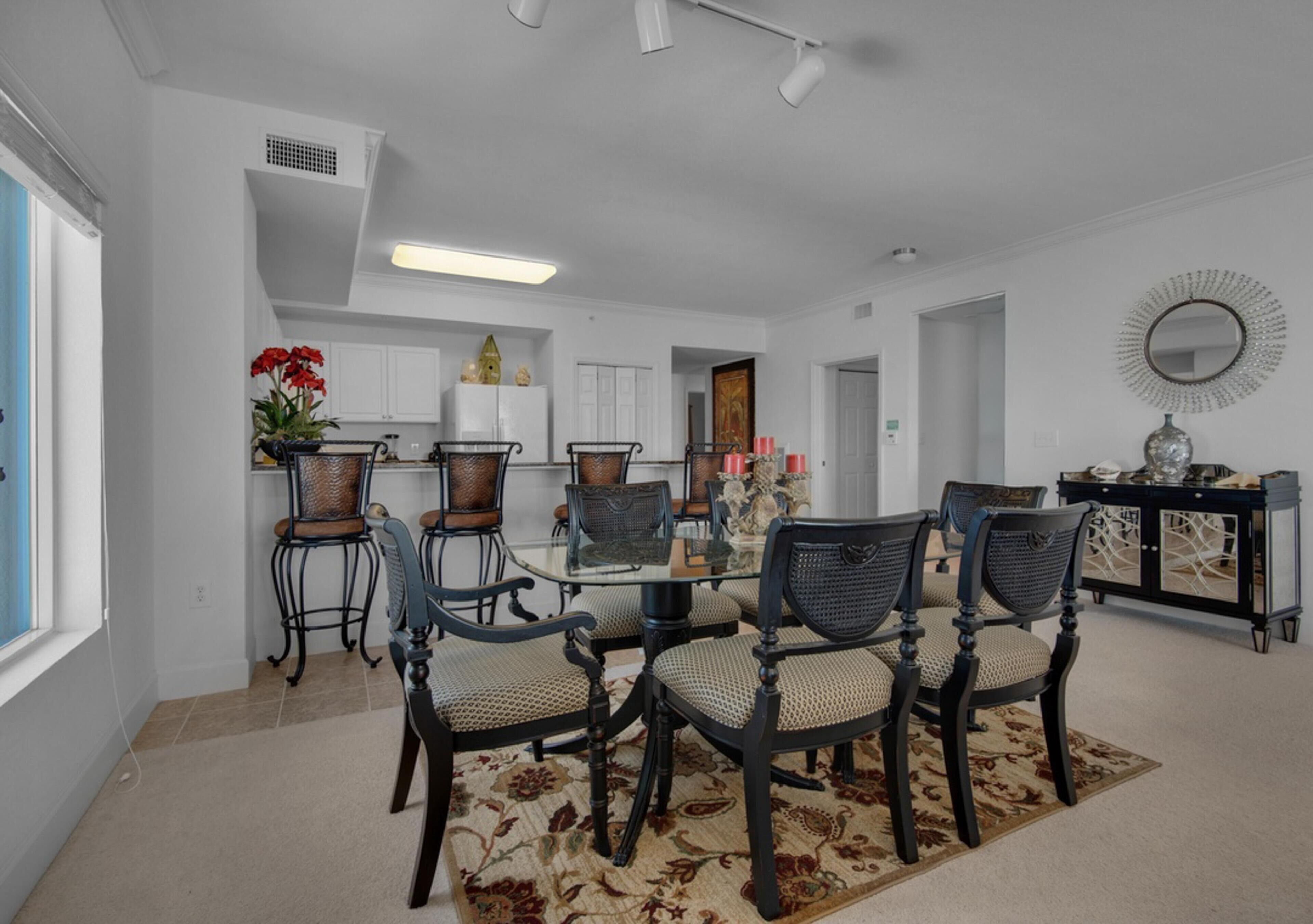 Un comedor con una mesa de vidrio rodeada de sillas, una cocina al fondo y un espejo decorativo en la pared.