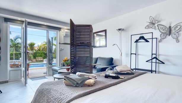 Une chambre d’hôtel moderne comprenant un lit, un coin salon avec un fauteuil et une table, un balcon avec vue sur les palmiers et une grande fenêtre.