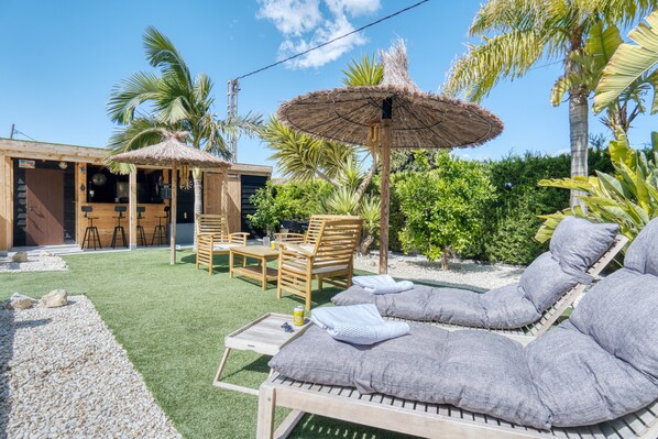 A patio area with a lounge chair, a wooden table, and a thatched umbrella.