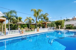 A swimming pool with lounge chairs and umbrellas, surrounded by palm trees and a white fence.