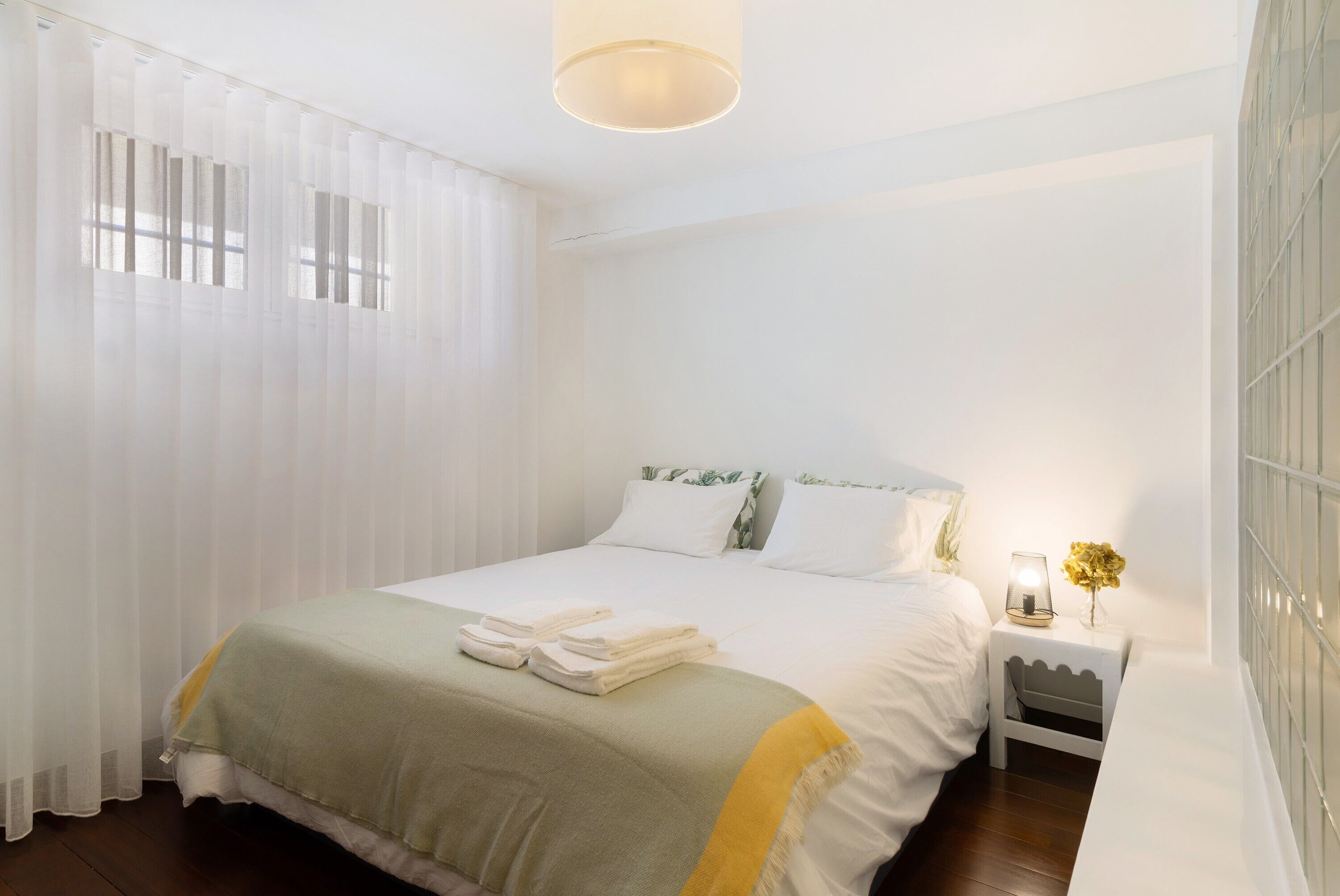 A neatly made bed with a green and white bedspread, a bedside table with a lamp and a vase of flowers, and sheer curtains allowing natural light.