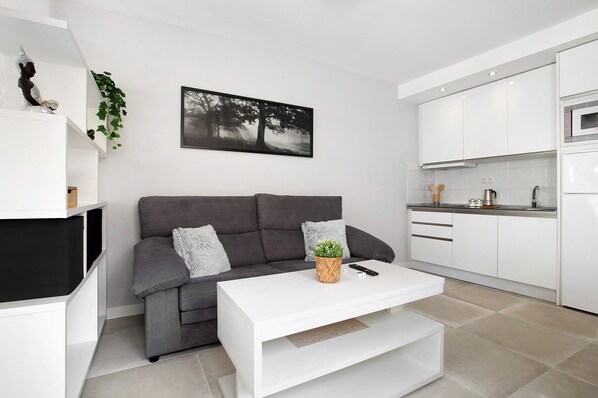Un salon moderne avec un canapé gris, une table basse blanche et un coin cuisine doté de meubles et d’appareils électroménagers blancs.